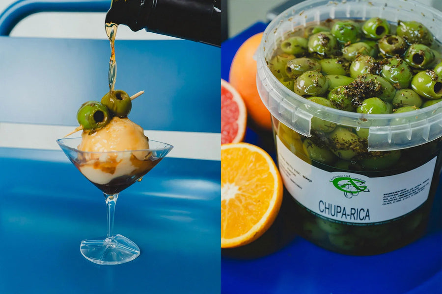Copa con una bola de helado de vermut y una brocheta de aceitunas, con el vermut sirviéndose por encima, junto a un cubo de aceitunas aliñadas y cítricos cortados