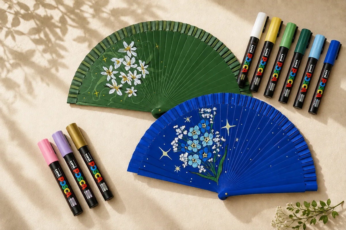 Dos abanicos pintados a mano, uno verde con margaritas y otro azul con nomeolvides, junto a los rotuladores Posca del taller