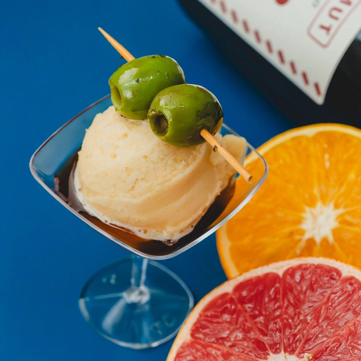 Copa con una bola de helado de vermut y una brocheta de aceitunas, junto a media naranja y medio pomelo sobre fondo azul