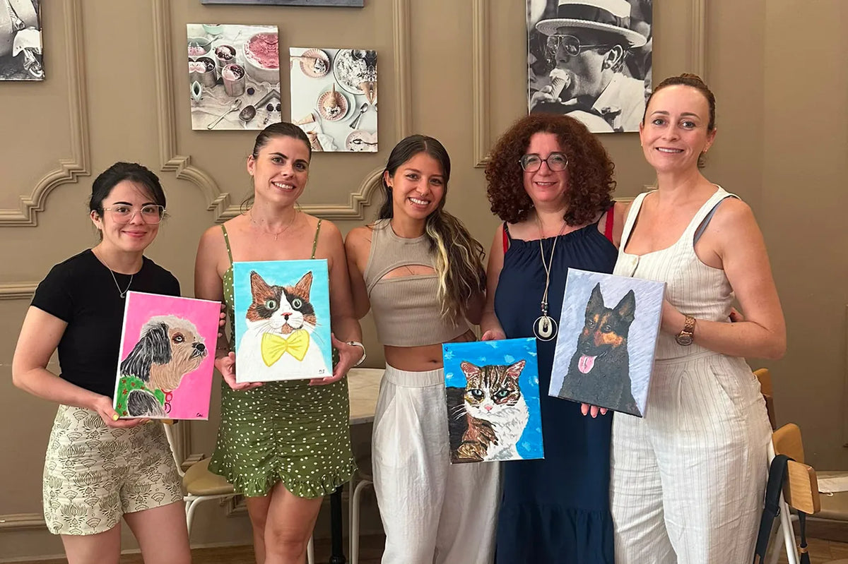 Cinco participantes del taller sostienen los retratos de gato y de perro que han pintado, de pie delante de la pared de la gelatería