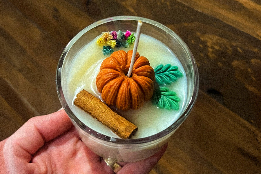 Vela de soja en tarro de cristal decorada con una calabaza de cera, hojas verdes, una rama de canela y flores secas, sostenida en la mano
