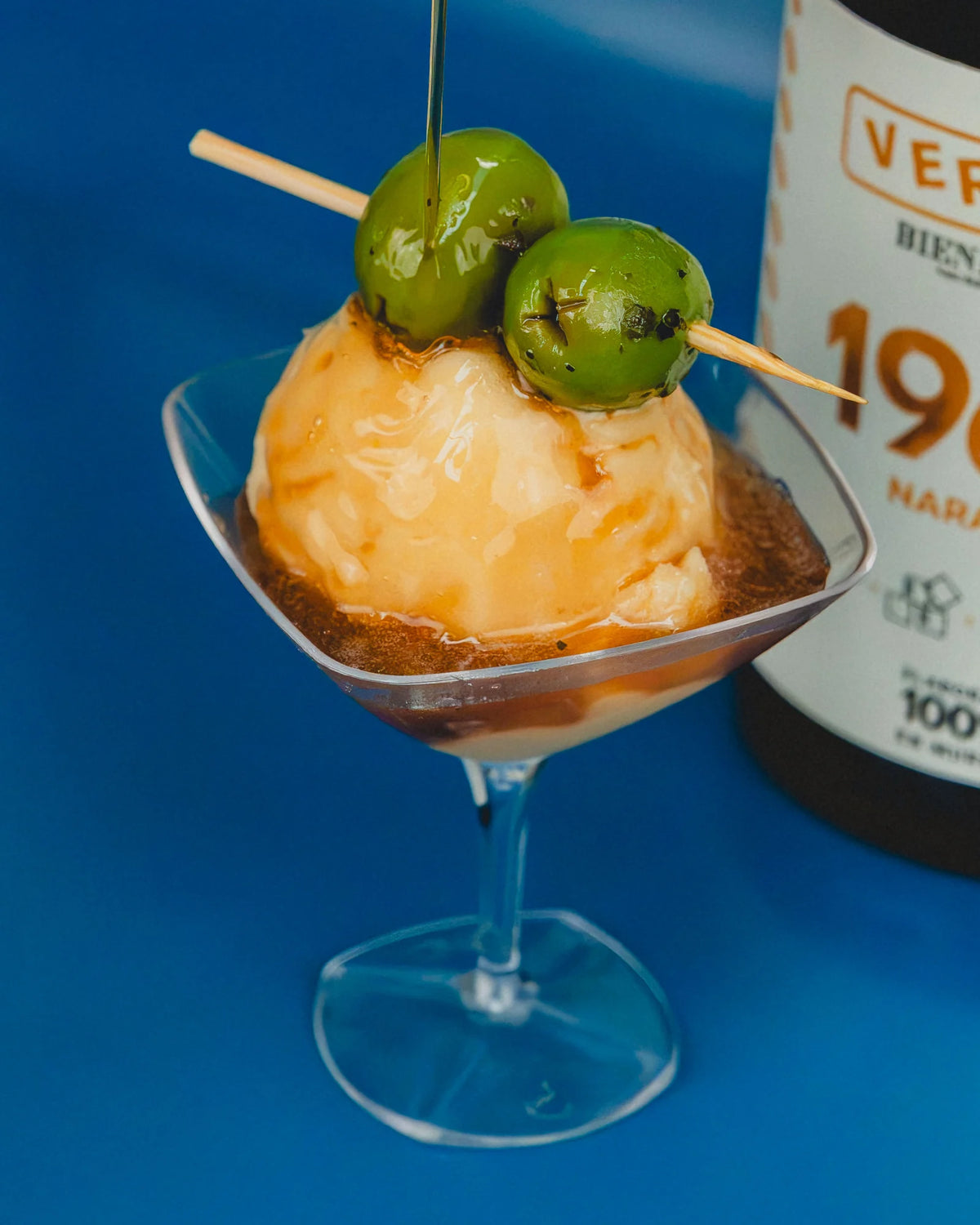 Copa con una bola de helado de vermut y su brocheta de aceitunas, con la botella de vermut Bienhablao 1968 detrás